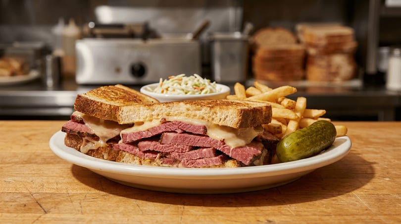 Grilled pastrami and cheese sandwich, fries, coleslaw, and a pickle on a plate in a diner.