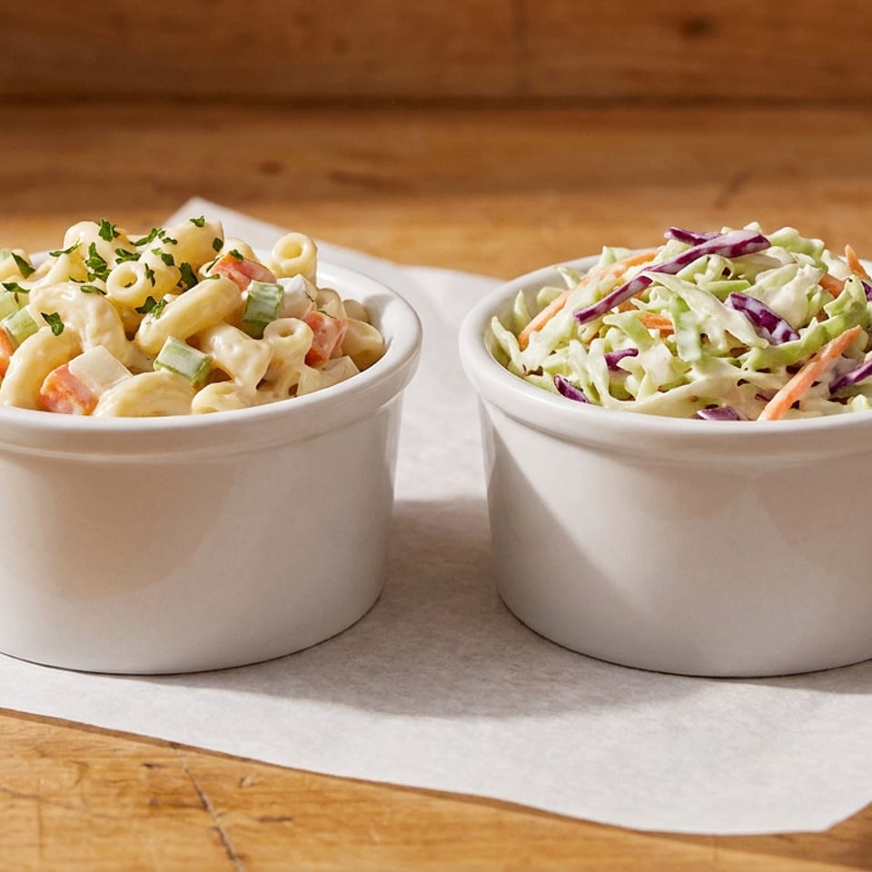 Macaroni salad and coleslaw in white bowls on parchment paper over a wooden table.