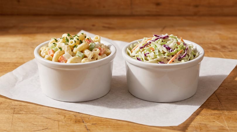 Macaroni salad and coleslaw in white bowls on parchment paper over a wooden table.