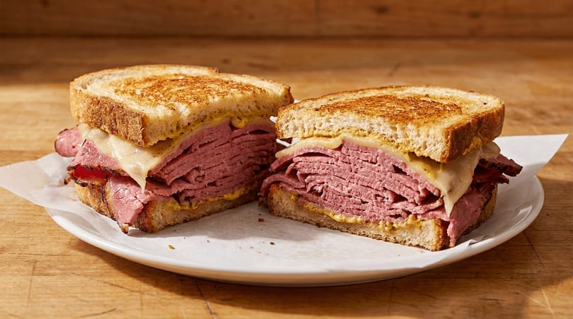 Toasted sandwich with sliced meat, melted cheese, and mustard, cut in half on a white plate.