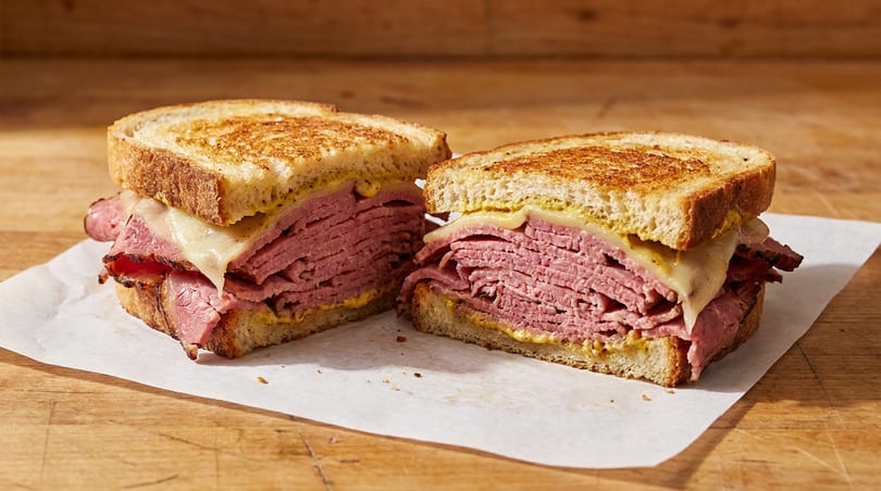Toasted sandwich cut in half, revealing layers of pastrami, melted cheese, and mustard on a wooden surface.