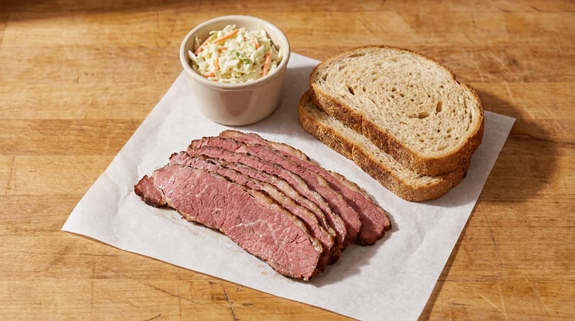 Deli-style sliced corned beef, rye bread, and coleslaw on a wooden surface.